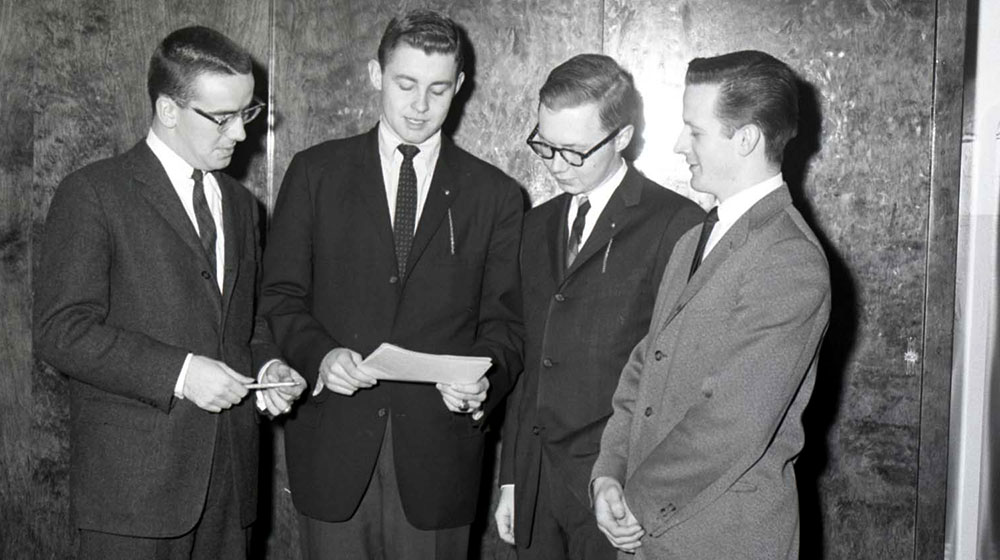 Bill Plante wearing a suit and tie, with three other young men all in suits and wearing ties, standing together looking down at a paper.