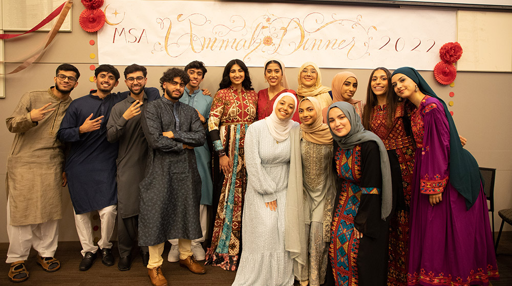 a group of students huddled together posing inside, Ummah Dinner is written on a poster in the back 