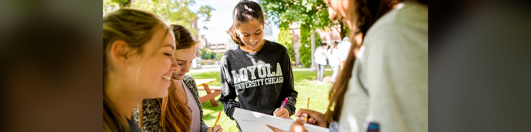 Several female students stand in a circle, outside on a sunny day with trees in bloom, each writing on a piece of paper. There is one student in focus, wearing a Loyola University Chicago shirt, smiling. Her dark hair is pulled back in a ponytail. Other students are slightly out of focus, but also smiling and talking.