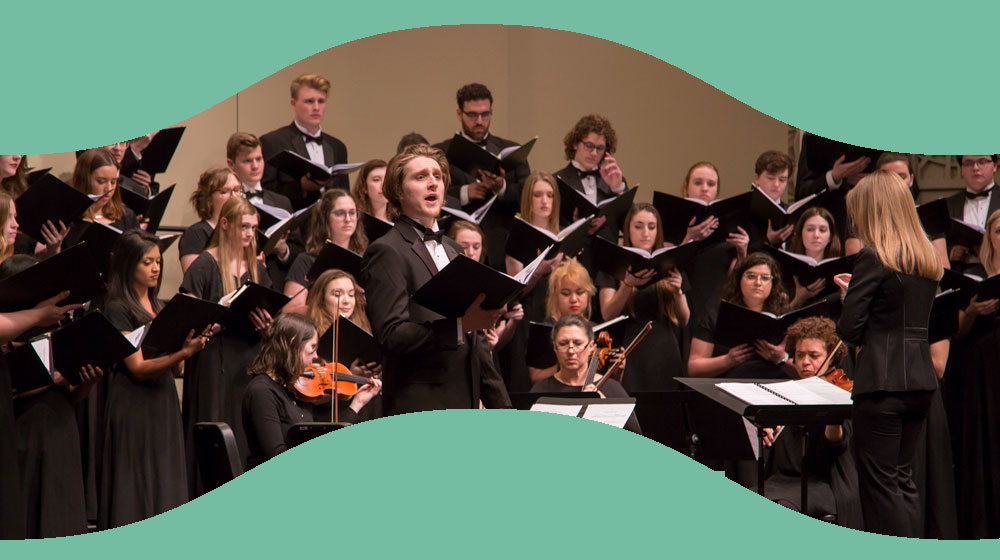 A group of musicians dressed in formal attire and seated like a orchestra. The conductor and a singing male soloist stand.