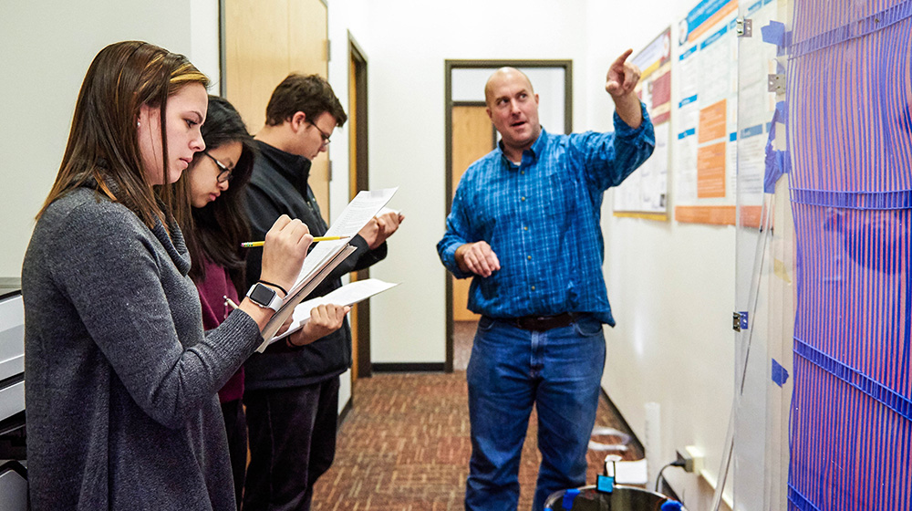 A group of ABET students take notes as they listen to an instructor dressed in a blue flannel shirt and jeans.