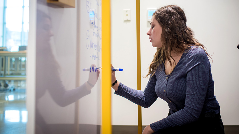 A young woman student with brown hair, writes on a whiteboard.