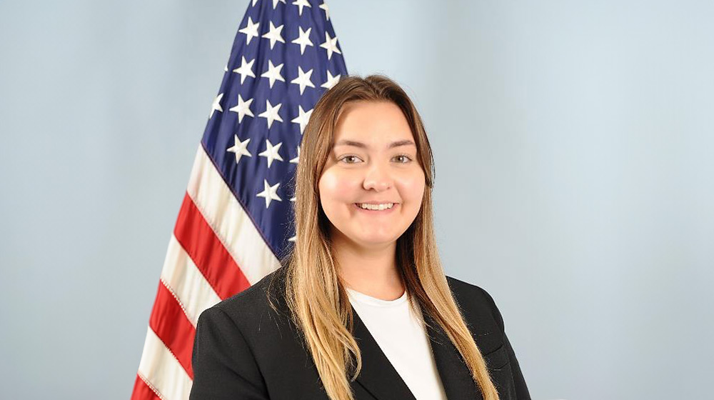 Headshot of a blond person in a black suit and white shirt; American flag in background