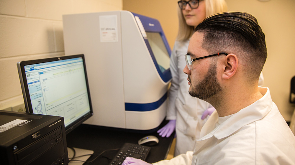 Two students looking at a computer screen near a piece of lab equipment.