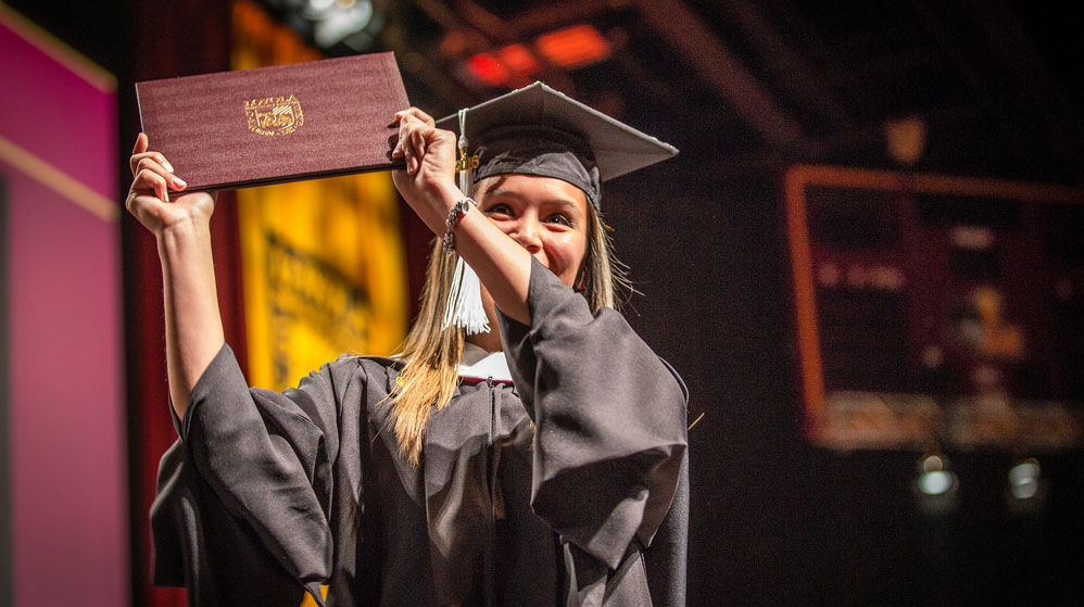 A woman in a graduation gown proudly holds her diploma, symbolizing her academic achievement and success.