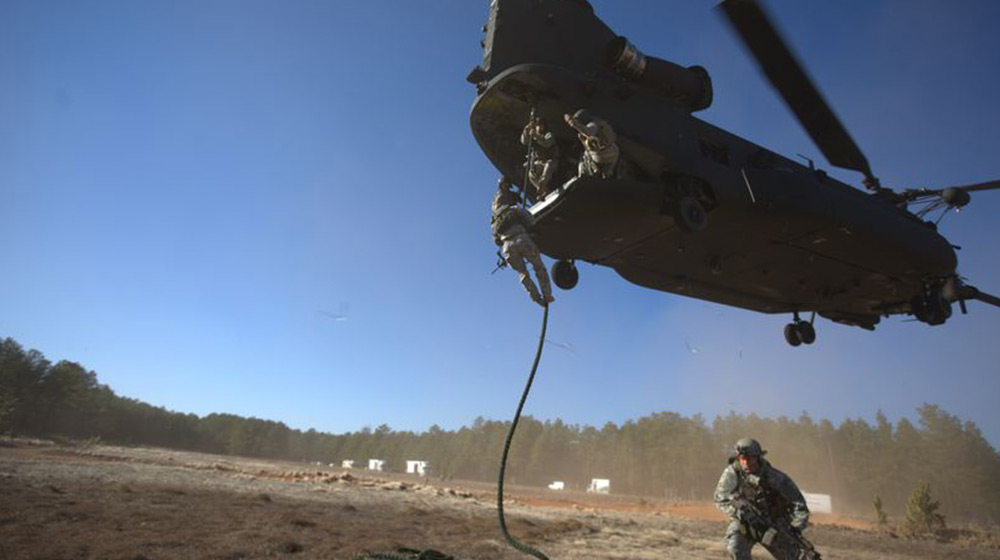 A military soldier rappels from an aircraft using a rope. Another trainee on the ground is looking ahead. They are in a clearing in a forest.