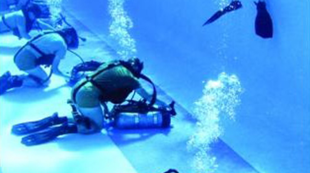 Military trainees are underwater in a pool with scuba gear attached to them. Large streams of air bubbles are seen coming from their heads as instructors watch their training.