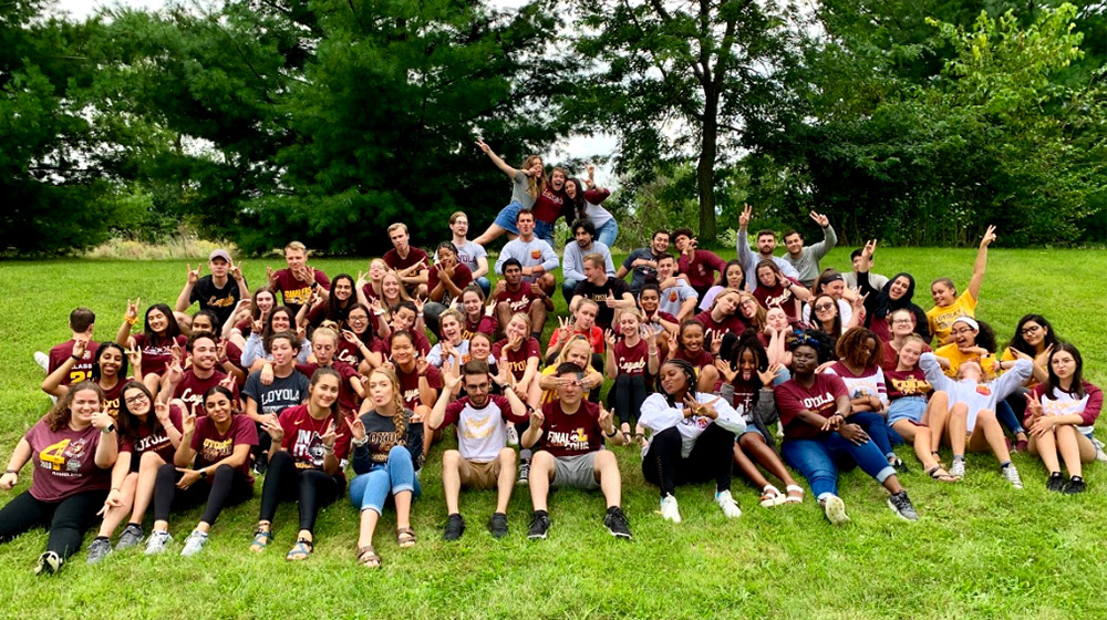 Dozens of Loyola students gather for a team photo on a grassy hill with a tree line behind them.