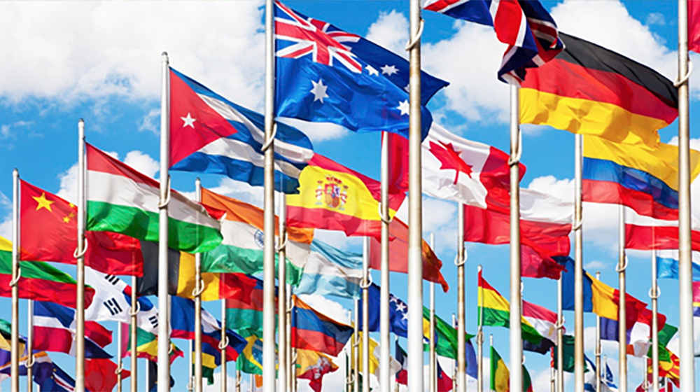 A large number of flags representing a wide range of countries wave against a blue sky filled with small, white, clouds.