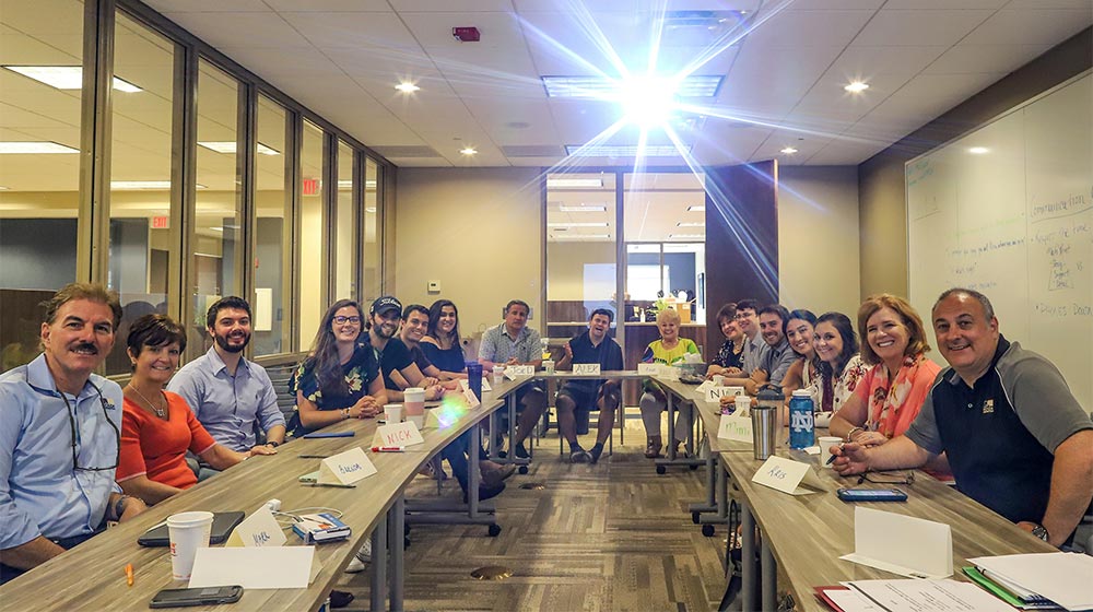 Seventeen people sit around  a U-shaped table looking at the camera smiling for a photo in a conference room with the overhead projector light on.