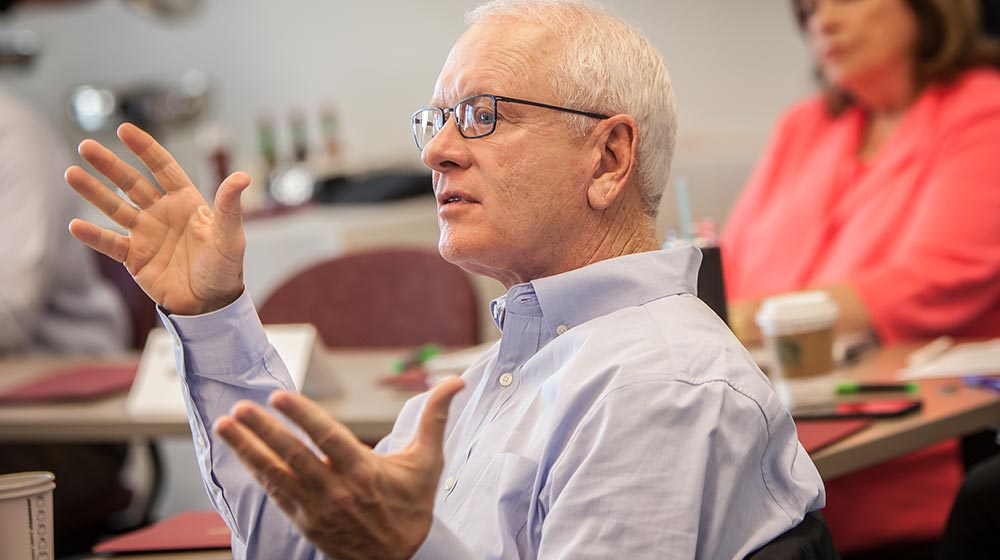 A man in profile is looking to the left with his arms up posing a question to a classroom. He has white hair, glasses, and a blue button down shirt.