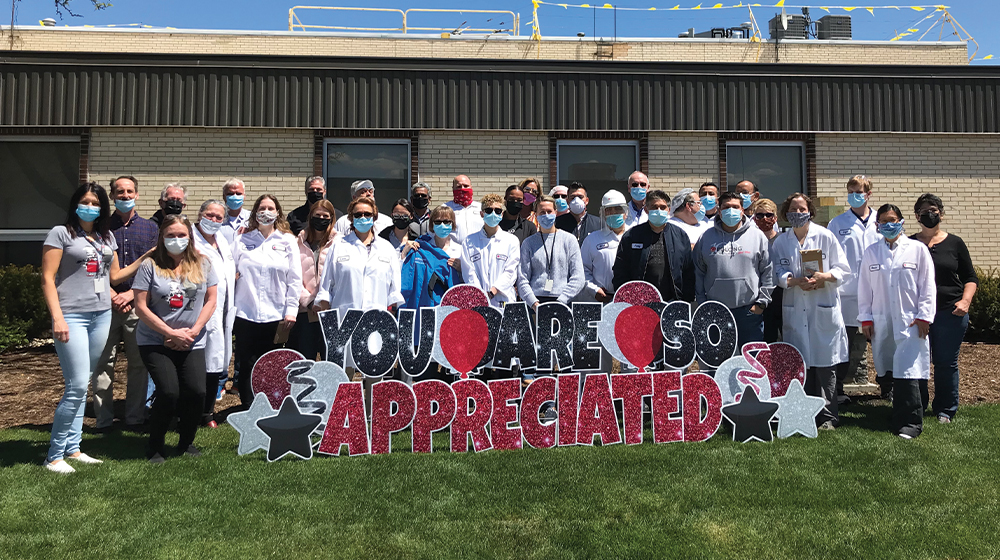 A large group of factory, research, and office workers in PPE masks pose in front of a building with a sign that reads 