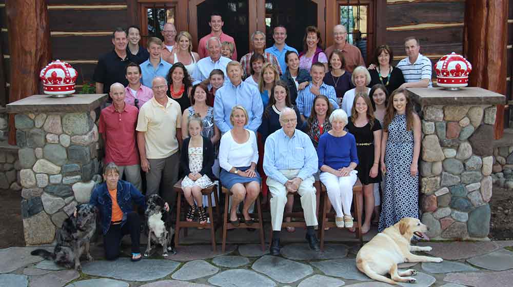 A photo of the Wall family with numerous members posed smiling at the camera in front of a wood and stone building. There are also 3 family dogs in the photo.