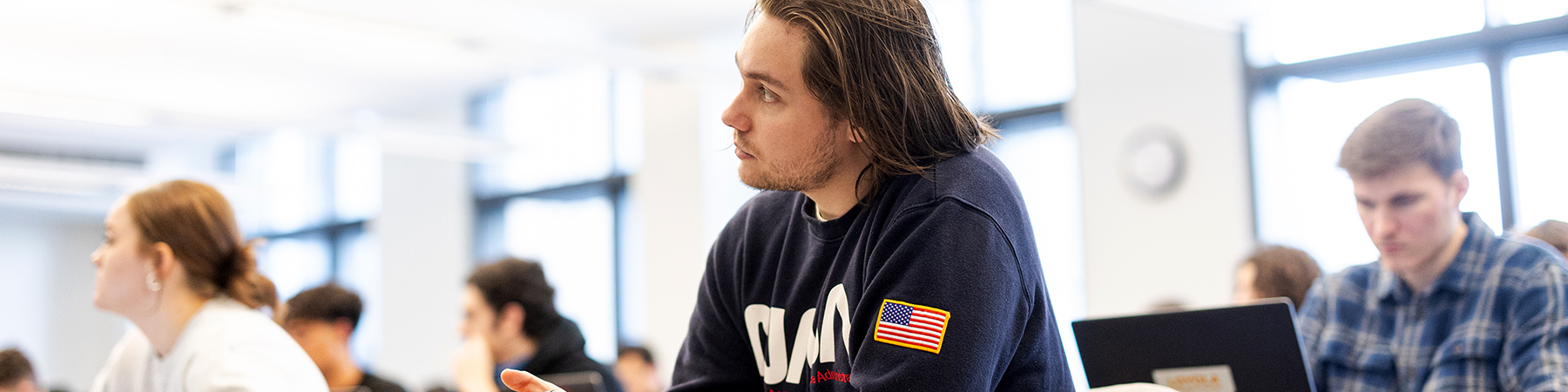 A young student with shoulder length brown hair listens intently to the instructor in a classroom with other students.. He wears a dark blue hoodie.