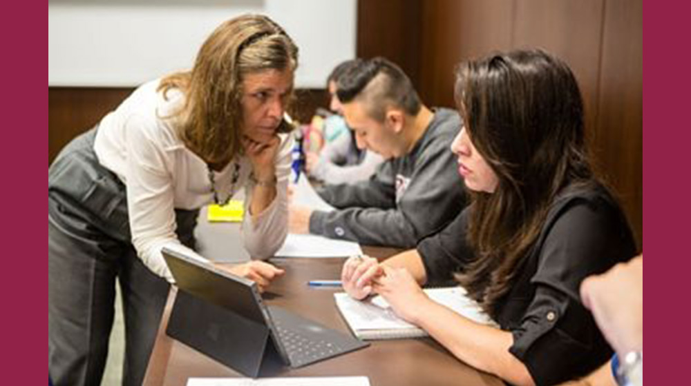 A professor leans on a table and listens to a student explain something on their laptop