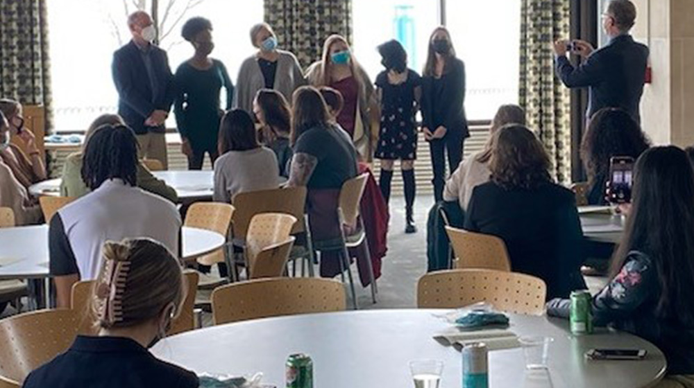 A large group of people gather in a room at large round tables and chairs for an award ceremony. Six individuals who won awards stand in front of ever