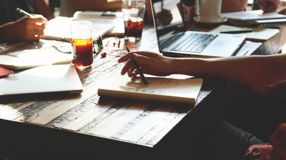 A group of people sitting around a table working with laptops, notebooks, and drinks. The scene suggests a collaborative meeting or study session in a casual setting. Sunlight filters through windows, casting shadows on the wooden table.