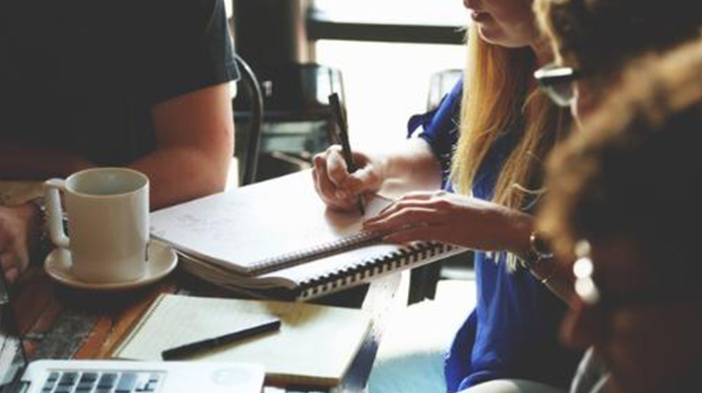 Four people gather at a table for a meeting. One person is taking notes in a notebook with a pen. There is a coffee cup and a laptop on the table.