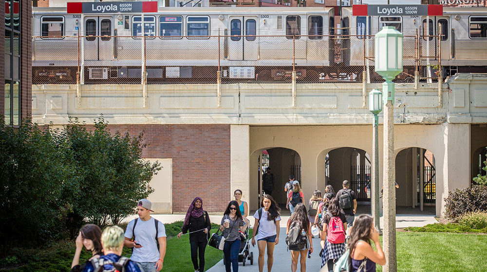 A group of people, primarily students, walk on a grassy path under a large elevated train track. An 