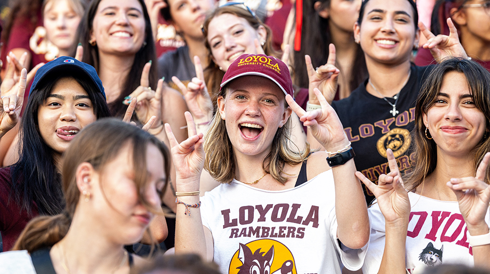 Several students with smiling faces, mostly young women, stand together on unseen bleachers wearing assorted short sleeve shirts an summer clothing.