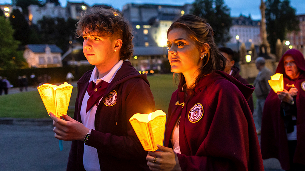 Nursing students walk together, carrying lanterns and wear maroon hooded capes.